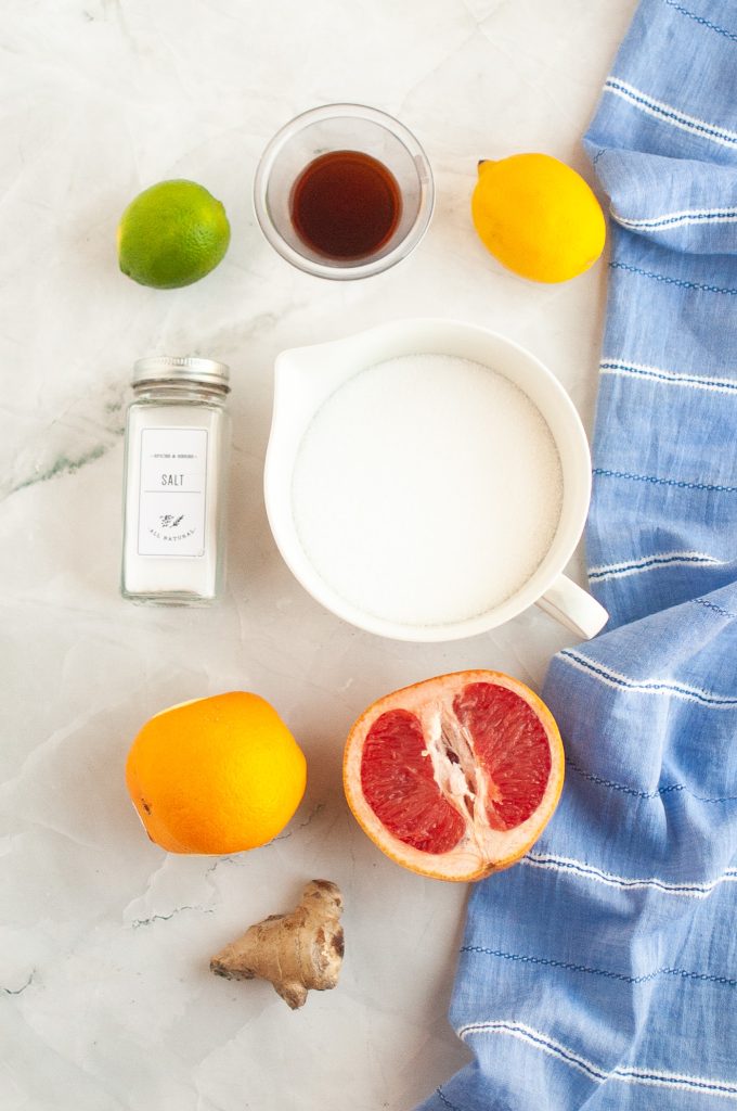 ingredients for small batch mixed citrus marmalade: A bowl of sugar, a halved grapefruit, an orange, a lemon, a lime, a ginger root, a bottle of salt, a small cup of liquid, and a blue cloth are arranged on a white marble surface.
