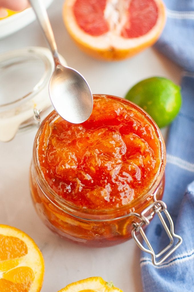 A spoon hovers over an open jar of mixed citrus marmalade, surrounded by halved citrus fruits and a blue cloth on a white surface.