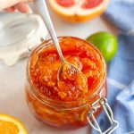 A hand holds a spoon scooping mixed citrus marmalade from a glass jar. Fresh citrus fruits, including orange halves, a grapefruit, and a lime, are visible on the table along with a blue cloth.