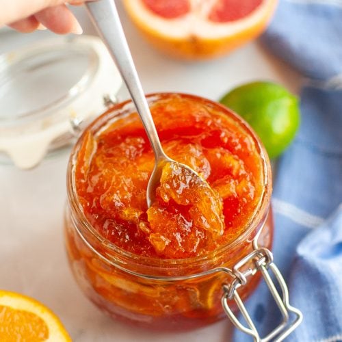 A hand holds a spoon scooping mixed citrus marmalade from a glass jar. Fresh citrus fruits, including orange halves, a grapefruit, and a lime, are visible on the table along with a blue cloth.