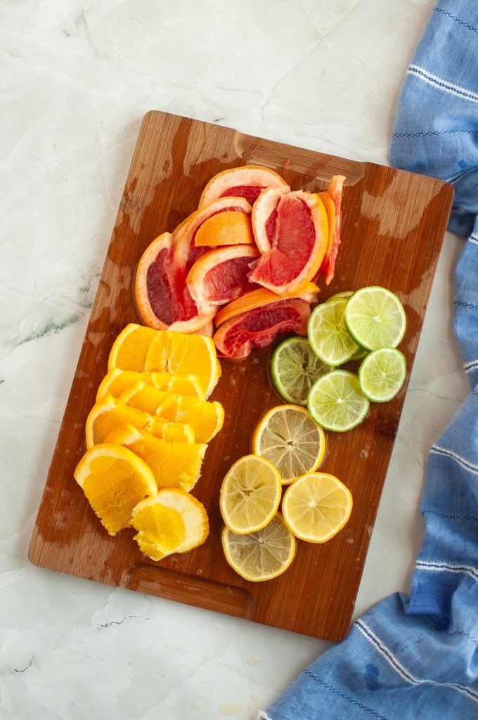 A wooden cutting board with neatly arranged slices of orange, grapefruit, lemon, and lime, next to a blue cloth on a light marble surface.
