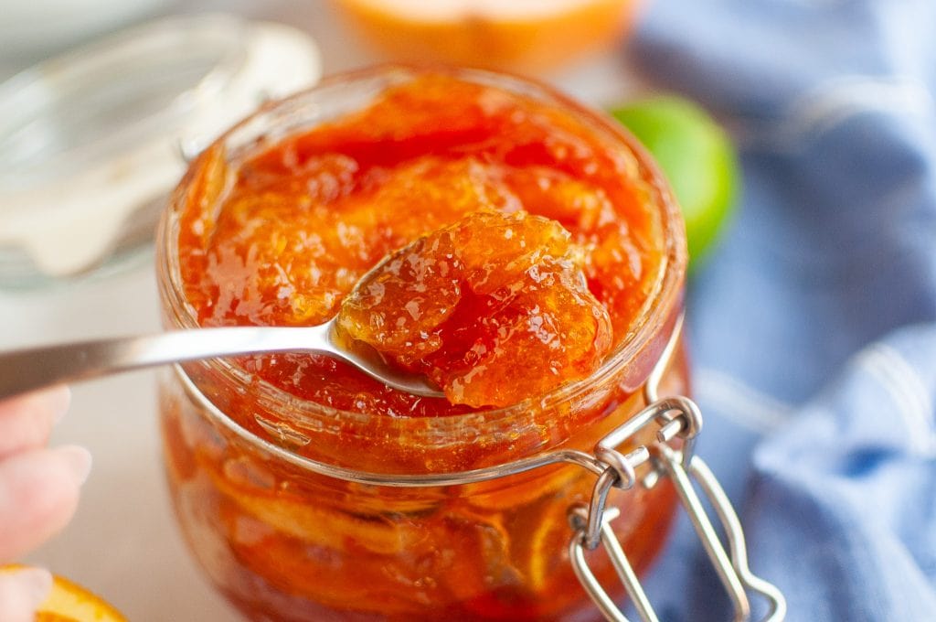 A close-up of a glass jar filled with mixed citrus marmalade. A spoon is lifting a portion of the marmalade from the jar. A blue cloth and some citrus fruit are blurred in the background.