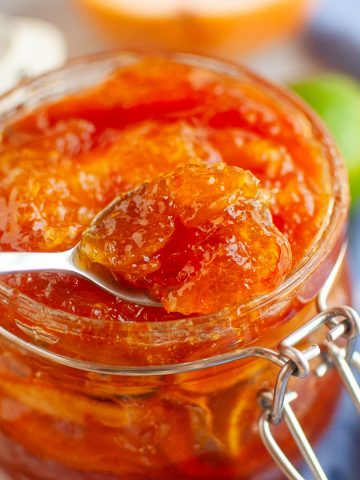 A close-up of a glass jar filled with mixed citrus marmalade. A spoon is lifting a portion of the marmalade from the jar. A blue cloth and some citrus fruit are blurred in the background.