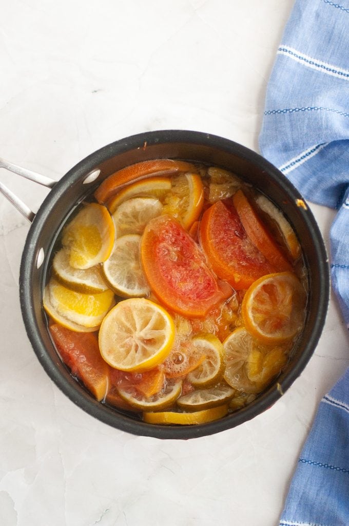 A saucepan filled with simmering slices of lemon, lime, orange, and grapefruit sits on a light marble surface next to a blue and white striped cloth.