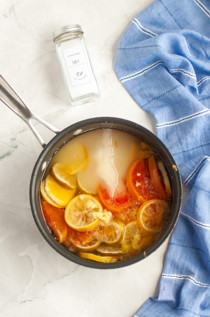 A saucepan contains simmering water with mixed citrus slices, and a large piece of ice. Nearby, a container of salt and a blue striped cloth are on a light marble surface.