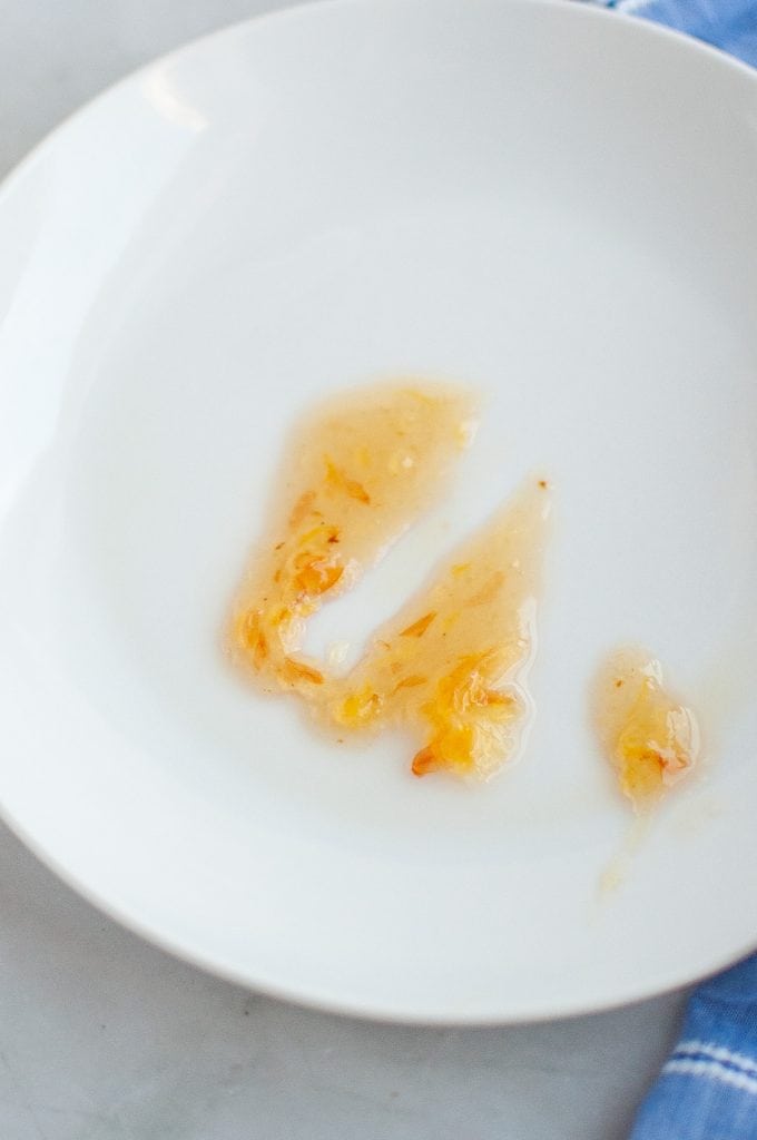A white plate with three smears of translucent orange sauce containing small chili flakes, placed on a light surface near a blue cloth.