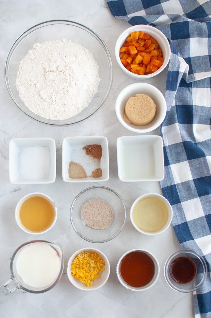 A top-down view of bowls containing flour, diced dried fruit, brown sugar, spices, salt, oil, yeast, non-dairymilk, citrus zest, agave,, vanilla, and other baking ingredients on a white surface with a blue checkered cloth.
