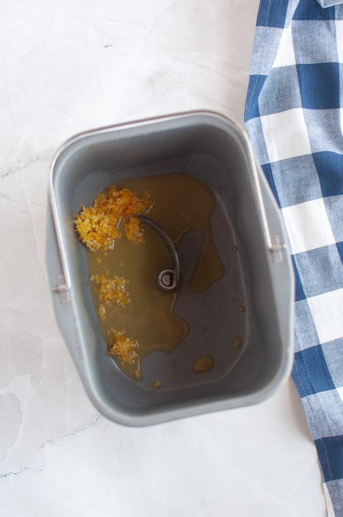 A metal bread machine container with fresh orange juice, zest, and a blue and white checkered cloth.