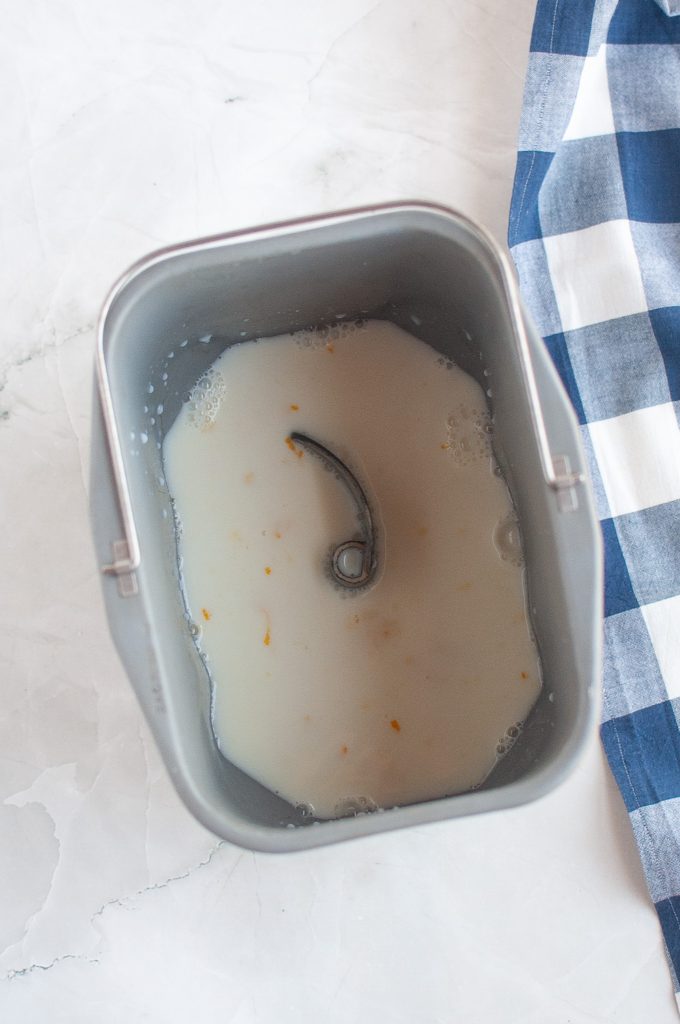 A bread machine pan with vegan milk and small orange zest pieces inside, placed on a white marble surface next to a blue and white checkered cloth.