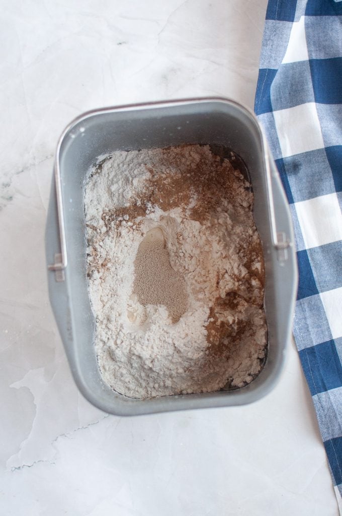 A bread machine pan filled with flour, yeast, and other dry ingredients sits on a white surface next to a blue and white checkered cloth.