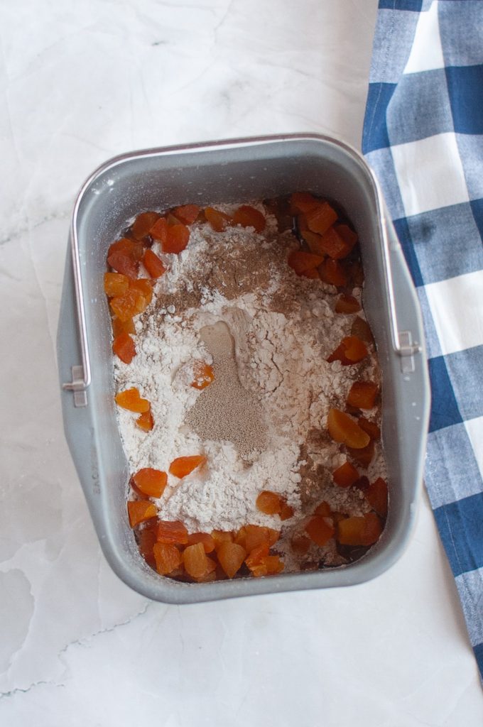 A bread machine pan containing flour, dried apricot pieces, and yeast, sits on a white marble surface next to a blue and white checkered cloth.