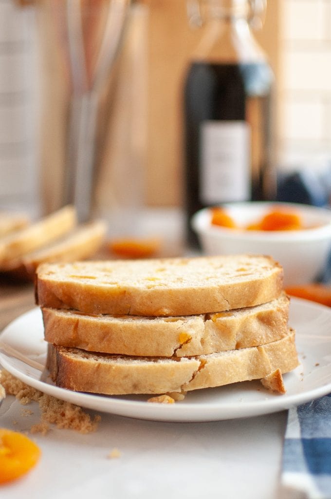 Three slices of bread machine apricot bread with apricot pieces are stacked on a white plate, with dried apricots and a bottle in the blurred background on a kitchen counter.