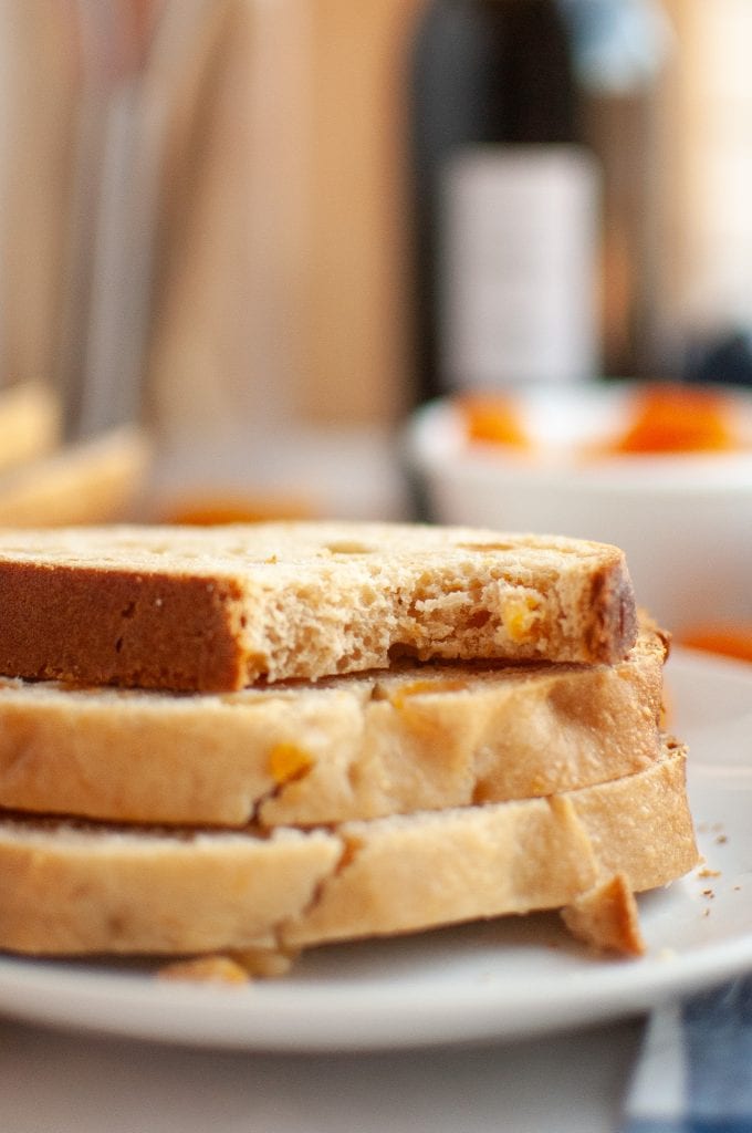 Three slices of bread machine apricot bread are stacked on a white plate, with the top slice partially eaten. The background is softly blurred, showing kitchen items and a bowl.