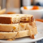 Three slices of vegan bread machine bread stacked on a white plate with a bite taken from the top slice; a blurred bowl with orange pieces is visible in the background.