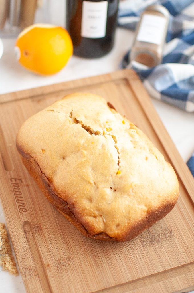 A golden-brown loaf of bread machine bread rests on a wooden cutting board. In the background, there is a whole orange, a bottle, a spice jar, and a blue-and-white cloth napkin.