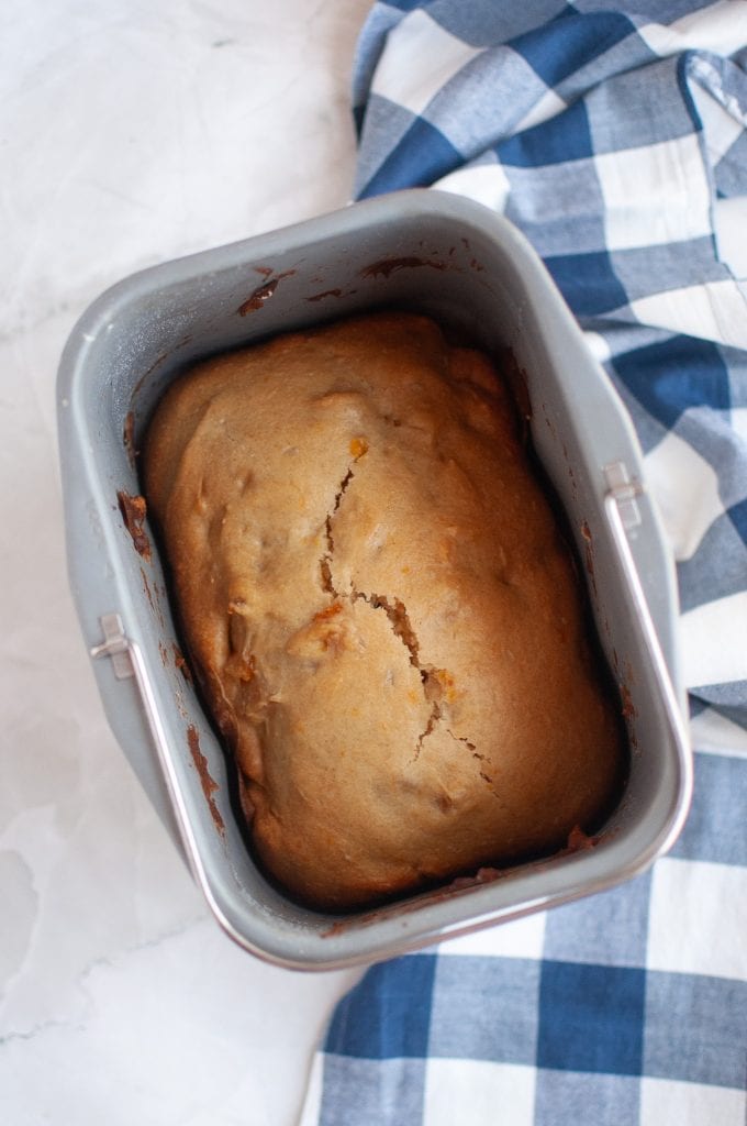 A loaf of bread with a cracked top sits in a bread machine pan next to a blue and white checkered cloth.