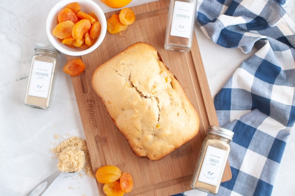 A loaf of bread machine apricot bread sits on a wooden cutting board, surrounded by dried apricots, ground cardamom, cinnamon, and ginger jars. A blue and white checkered cloth is nearby.