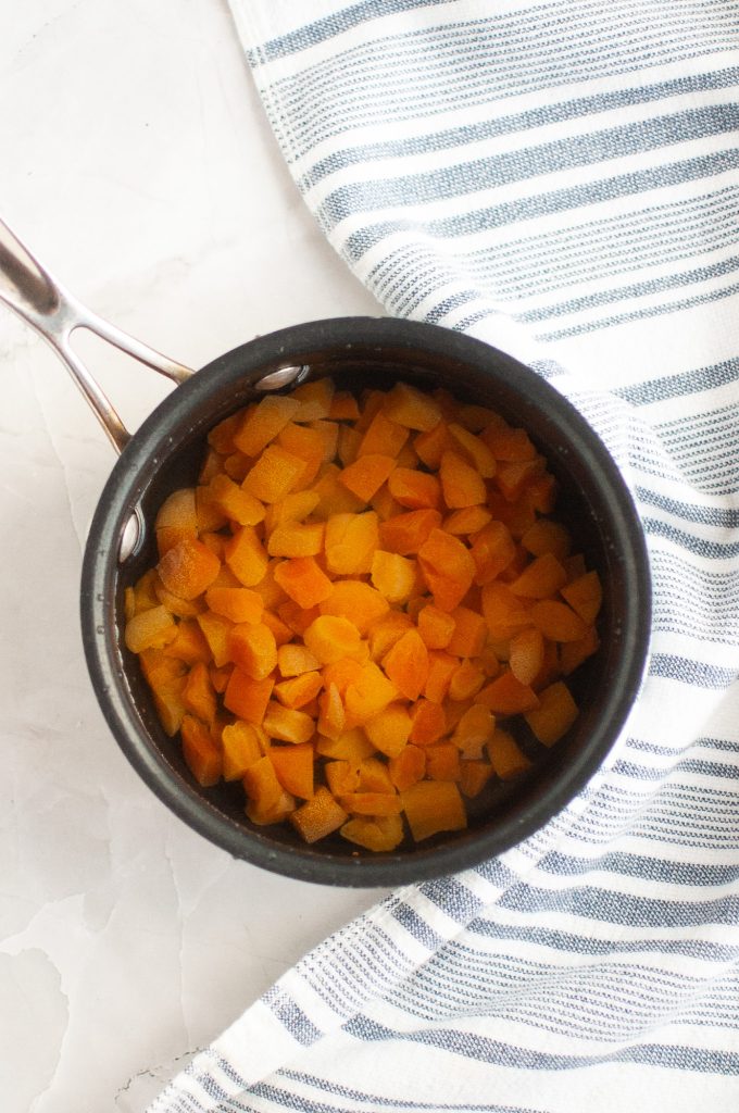 A small black saucepan filled with chopped dried apricots sits on a white marble surface next to a blue and white striped kitchen towel.