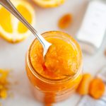 A spoon scoops bright apricot orange ginger jam from a glass jar. Sliced oranges, dried apricots, and a jar of spices are visible in the blurred background on a white surface.