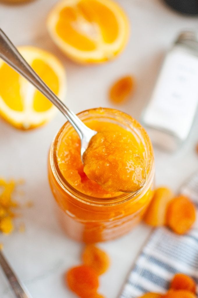 A spoon scoops bright apricot orange ginger jam from a glass jar. Sliced oranges, dried apricots, and a jar of spices are visible in the blurred background on a white surface.