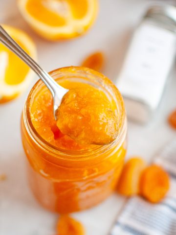 A spoon lifts apricot jam from a glass jar. Around it are halved oranges, dried apricots, citrus zest, a striped towel, and a spice jar on a white surface.