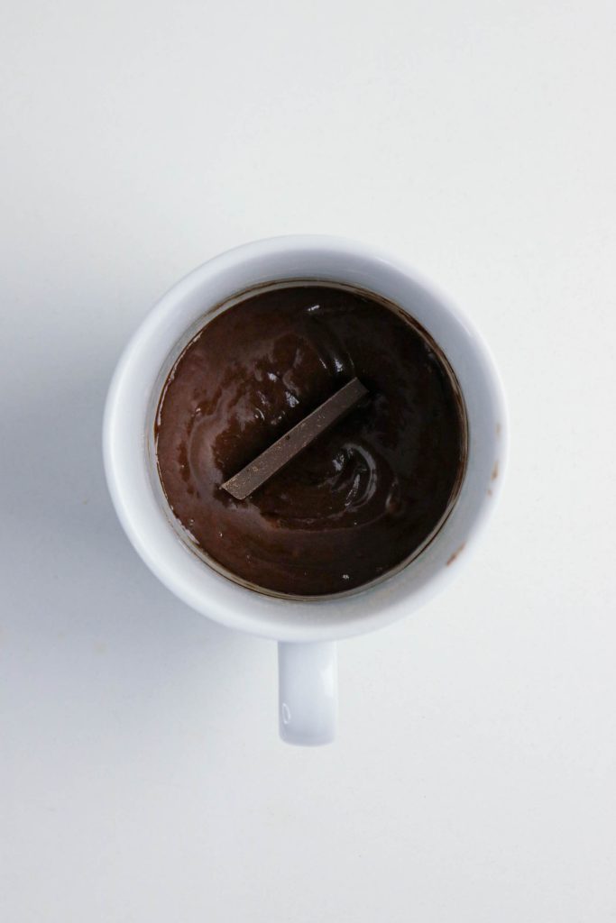 A white mug filled with thick, melted chocolate and a piece of vegan chocolate bar placed on top, viewed from above on a plain white background.