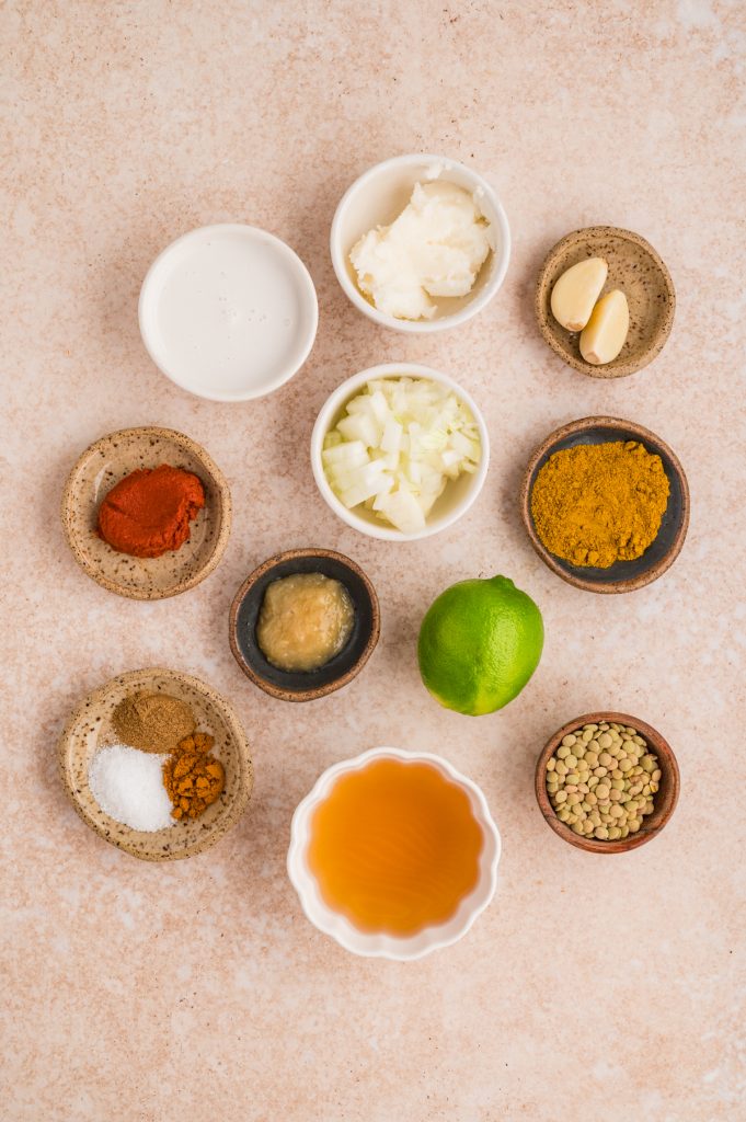 A flat lay of small bowls containing various ingredients for coconut curry lentil: chopped onions, curry powder, paprika, garlic cloves, lime, coconut milk, salt, garlic paste, a spice mix, brown lentils, and a bowl of vegetable broth.