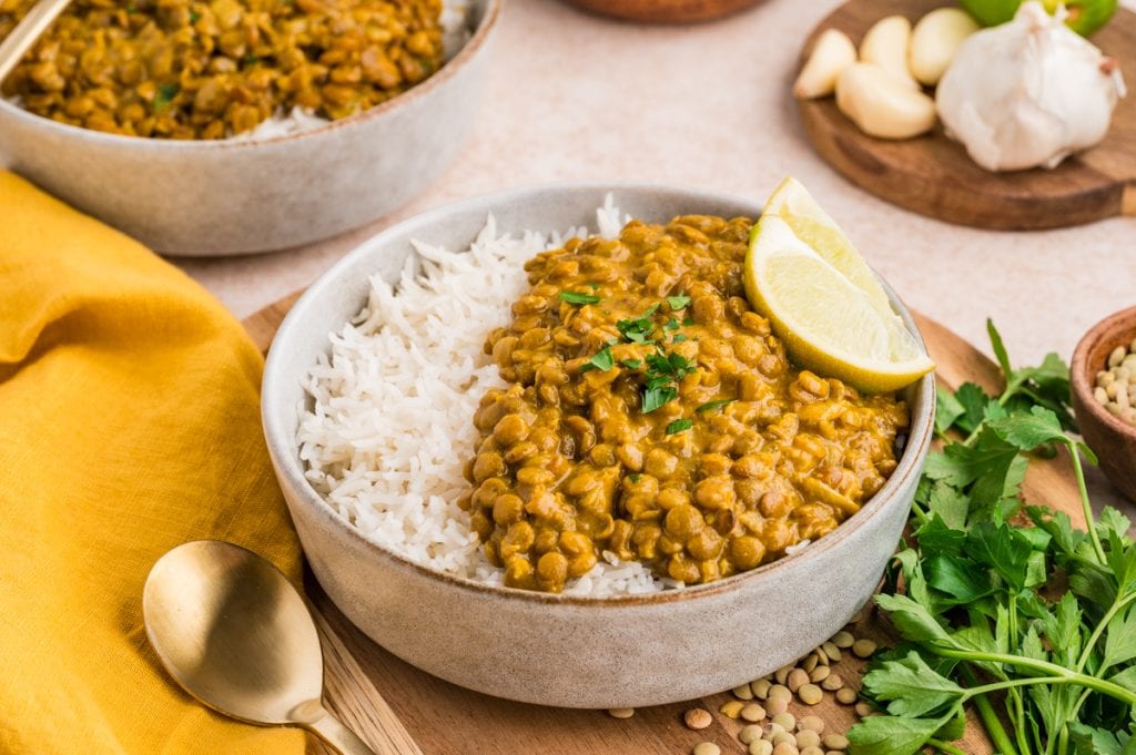 A bowl of cooked white rice topped with creamy coconut lentil curry, garnished with chopped herbs and two lemon wedges. Fresh parsley, garlic, lentils, and a gold spoon are nearby on the table.