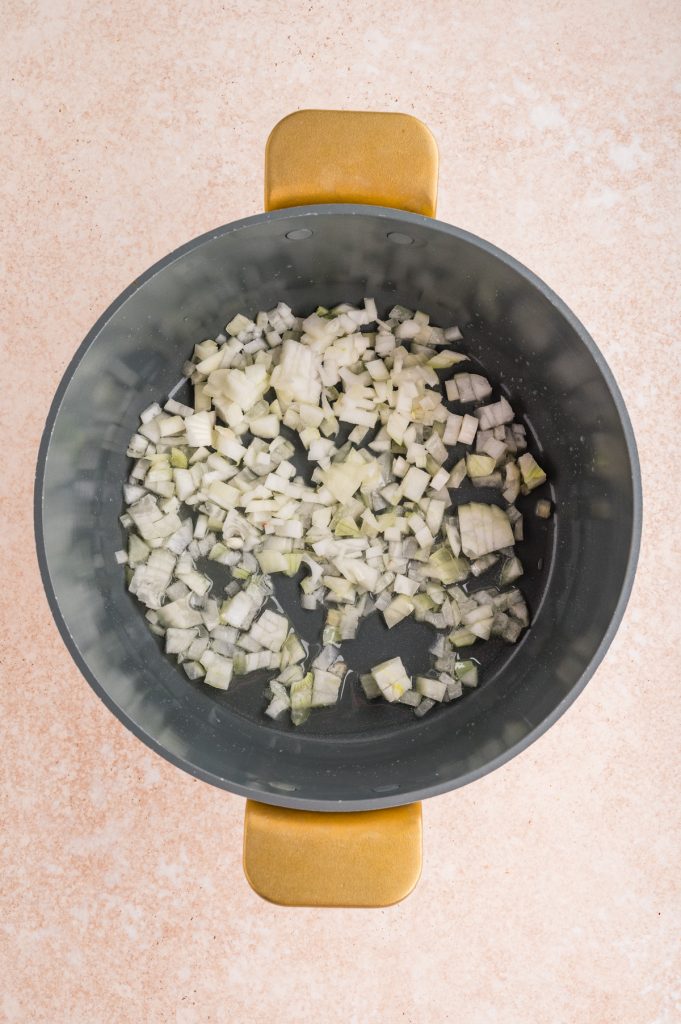 Diced onions sautéing in a large black pot with golden handles, placed on a light-colored countertop.
