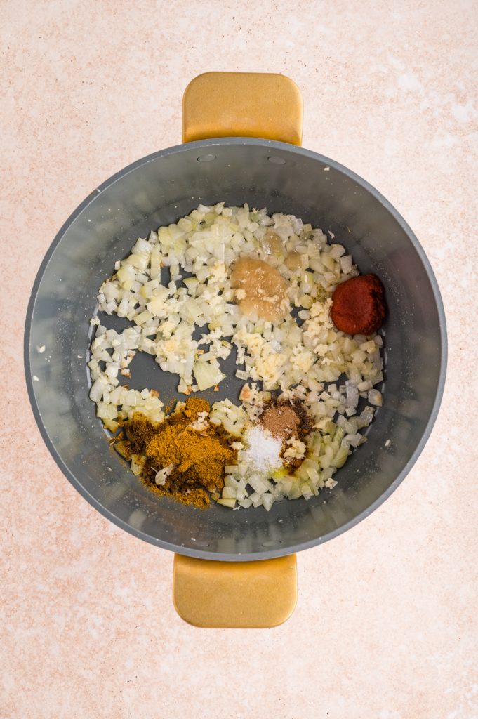 Chopped onions sautéing in a pot with assorted spices including curry powder, salt, ground ginger, viewed from above on a light-colored countertop.