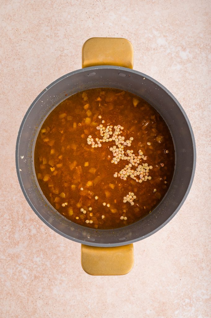 A top-down view of a pot filled with a vegetable broth containing chopped vegetables, curry spices, and small uncooked brown lentils, placed on a light-colored surface.