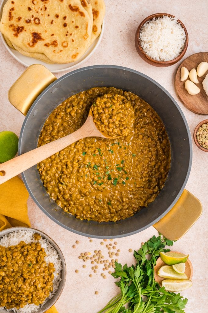 A pot of cooked coconut lentil curry with a wooden spoon, surrounded by rice, naan bread, garlic cloves, fresh parsley, lime wedges, and uncooked lentils on a light countertop.