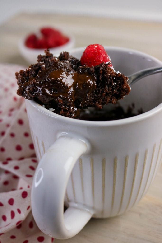 A spoonful of gooey molten chocolate mug lava cake topped with a raspberry is lifted from a white mug, with more raspberries in a small bowl in the background. A red and white polka dot cloth is beside the mug.