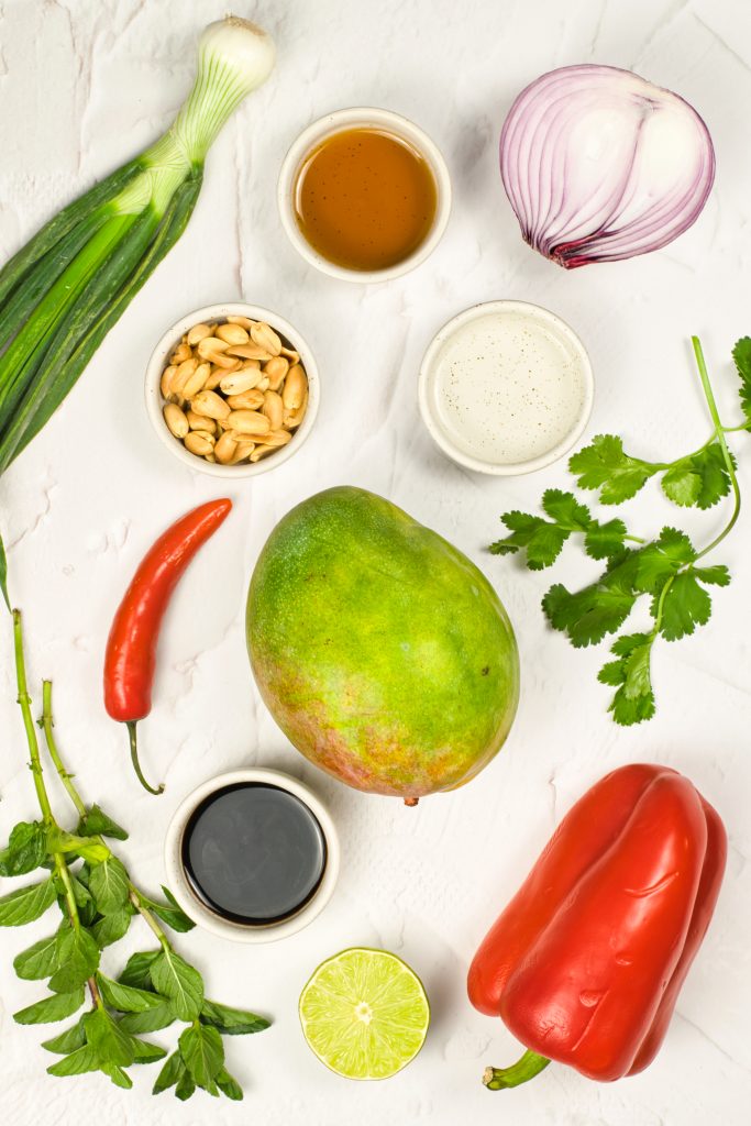 A flat lay of fresh ingredients on a white surface, including a whole mango, red bell pepper, chili, lime half, green onion, red onion, peanuts, cilantro, parsley, and small bowls of soy sauce, maple syrup, and lime juice.