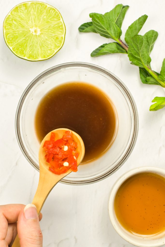 A hand holds a wooden spoon with chopped red chili over a bowl of brown sauce. Nearby are a halved lime, fresh mint leaves, and a small bowl of golden liquid, all on a white surface.