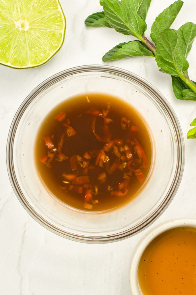 A glass bowl filled with a brown dressing containing chopped red chili and garlic sits on a white surface. Nearby are a half lime, fresh mint leaves, and a small bowl of golden liquid.