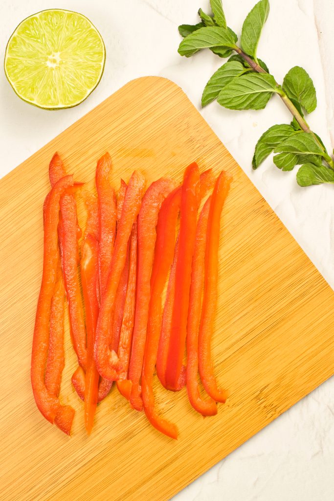 Thin slices of red bell pepper are arranged on a wooden cutting board. A halved lime and a sprig of fresh mint are placed nearby on a white surface.