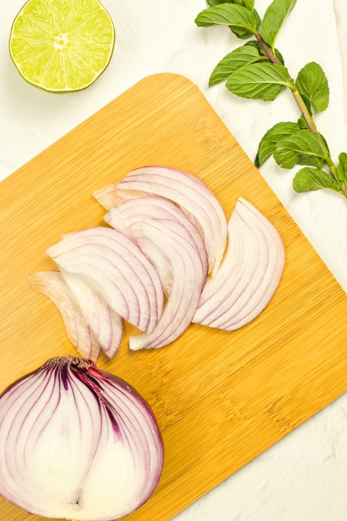 A halved red onion with several thin slices sits on a wooden cutting board. A sprig of mint and a halved lime are nearby on a white surface.