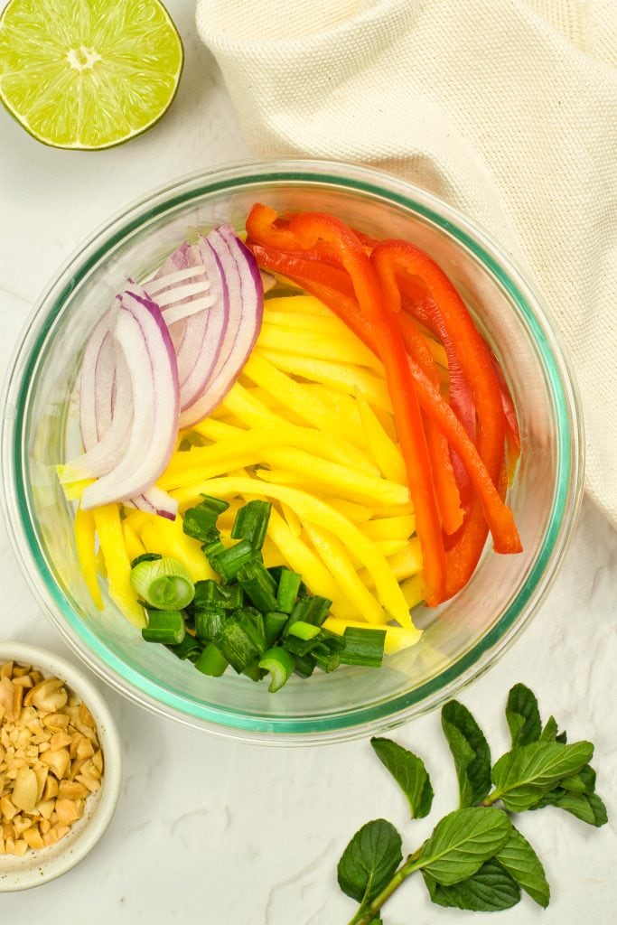 A glass bowl contains sliced red bell pepper, yellow mango, red onion, and green onion, arranged in sections. Surrounding the bowl are chopped peanuts, mint leaves, half a lime, and a beige cloth.