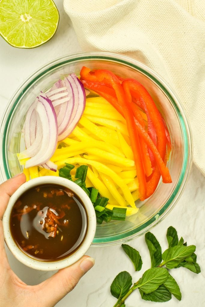 A glass bowl with sliced red bell pepper, yellow mango, red onion, and green onions. A hand holds a small bowl of soy dressing above, with mint leaves, a lime half, and a beige cloth nearby.