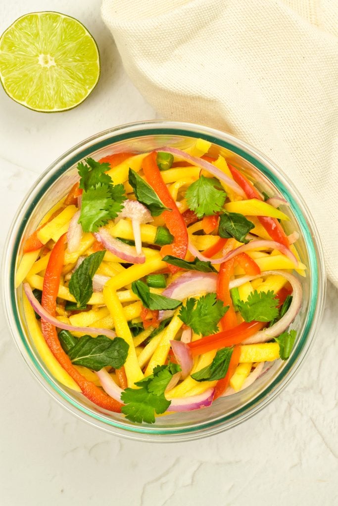 A glass bowl filled with a colorful mango salad containing sliced mango, red bell pepper, red onion, cilantro, and basil. A halved lime and a beige cloth are beside the bowl on a light surface.