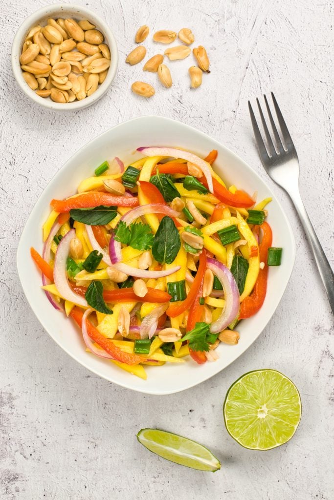A bowl of colorful salad with sliced red bell peppers, red onions, mango, green onions, cilantro, and peanuts, placed on a light surface with a fork, lime halves, and a small bowl of peanuts nearby.