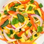 A colorful salad with sliced mango, red bell pepper, red onion, green onions, cilantro, mint leaves, and peanuts on a white plate.