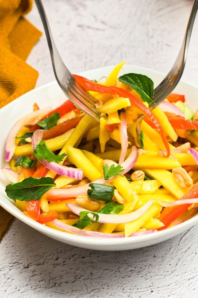 A bowl of colorful vegan mango salad with sliced red onions, red bell peppers, and fresh herbs, being tossed with serving utensils on a light surface.
