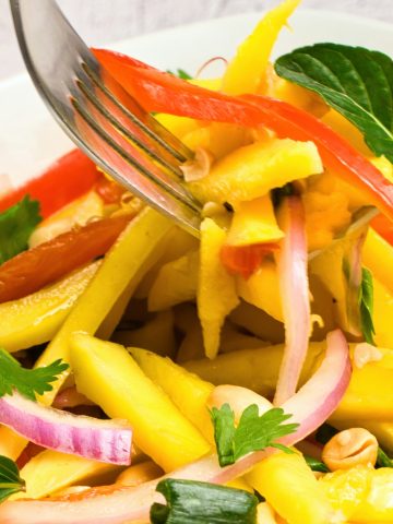 A close-up of a vibrant salad with sliced mango, red onion, red bell pepper, fresh herbs, peanuts, and mint leaves being tossed with two forks.