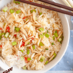 A bowl of pineapple coconut rice topped with sliced green onions and toasted slivered almonds, with chopsticks resting on top. Text reads: Vegan! Rice Cooker– Pineapple Coconut Rice. Veggie Fun Kitchen.