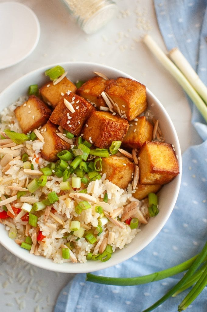 A white bowl filled with pan-fried tofu cubes, pineapple rice mixed with chopped green onions, slivered almonds, red bell peppers, and sesame seeds, with fresh green onions and a blue napkin nearby.
