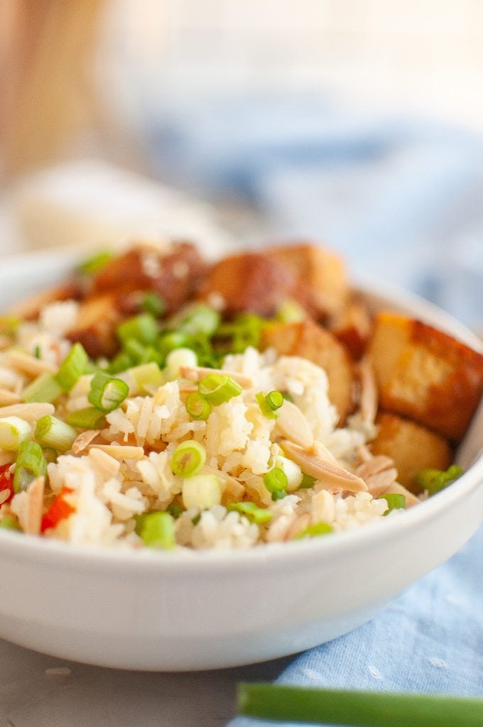 A close-up of a bowl filled with pineapple rice, slivered almonds, diced red peppers, green onions, and pieces of tofu, set on a light blue cloth.