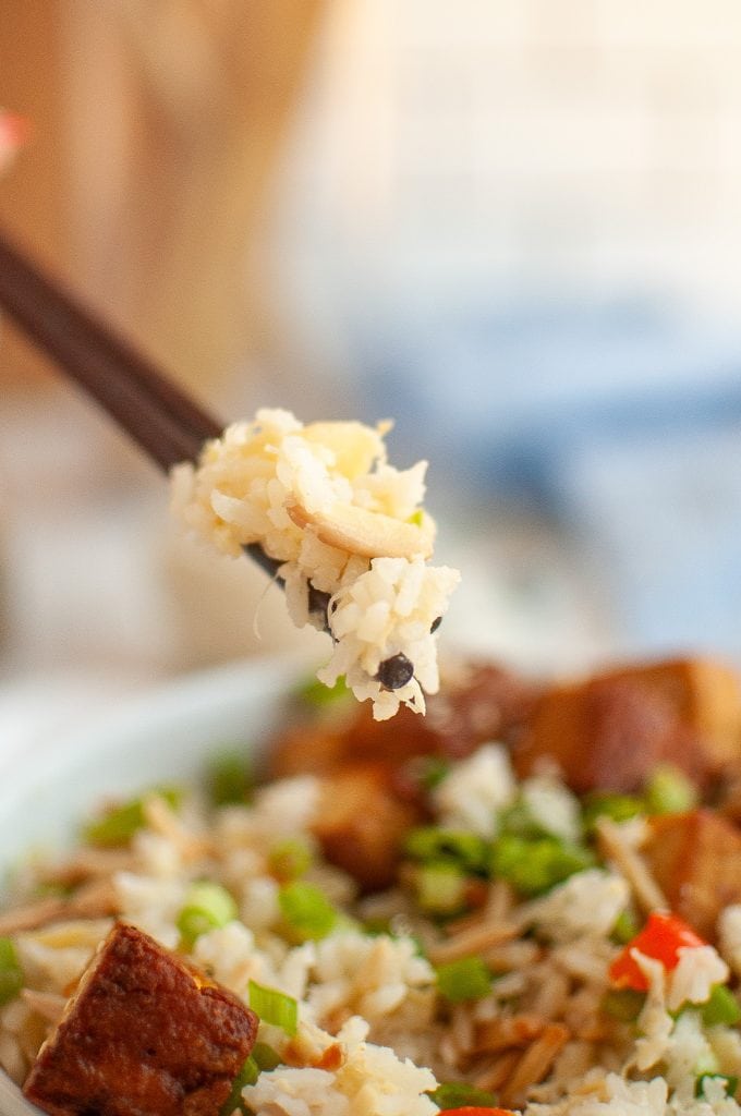 Close-up of chopsticks holding a bite of pineapple rice with vegetables and tofu, above a plate filled with more pineapple rice, tofu cubes, and chopped green onions.