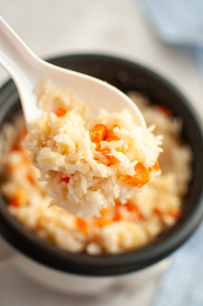 A close-up of a white plastic spoon holding a scoop of cooked pineapple rice mixed with diced red bell peppers, hovering over a bowl of the same rice mixture.